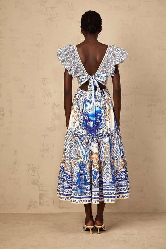A blue midi dress with a baroque print pattern featuring tiered layers worn by a woman standing in a neutral setting