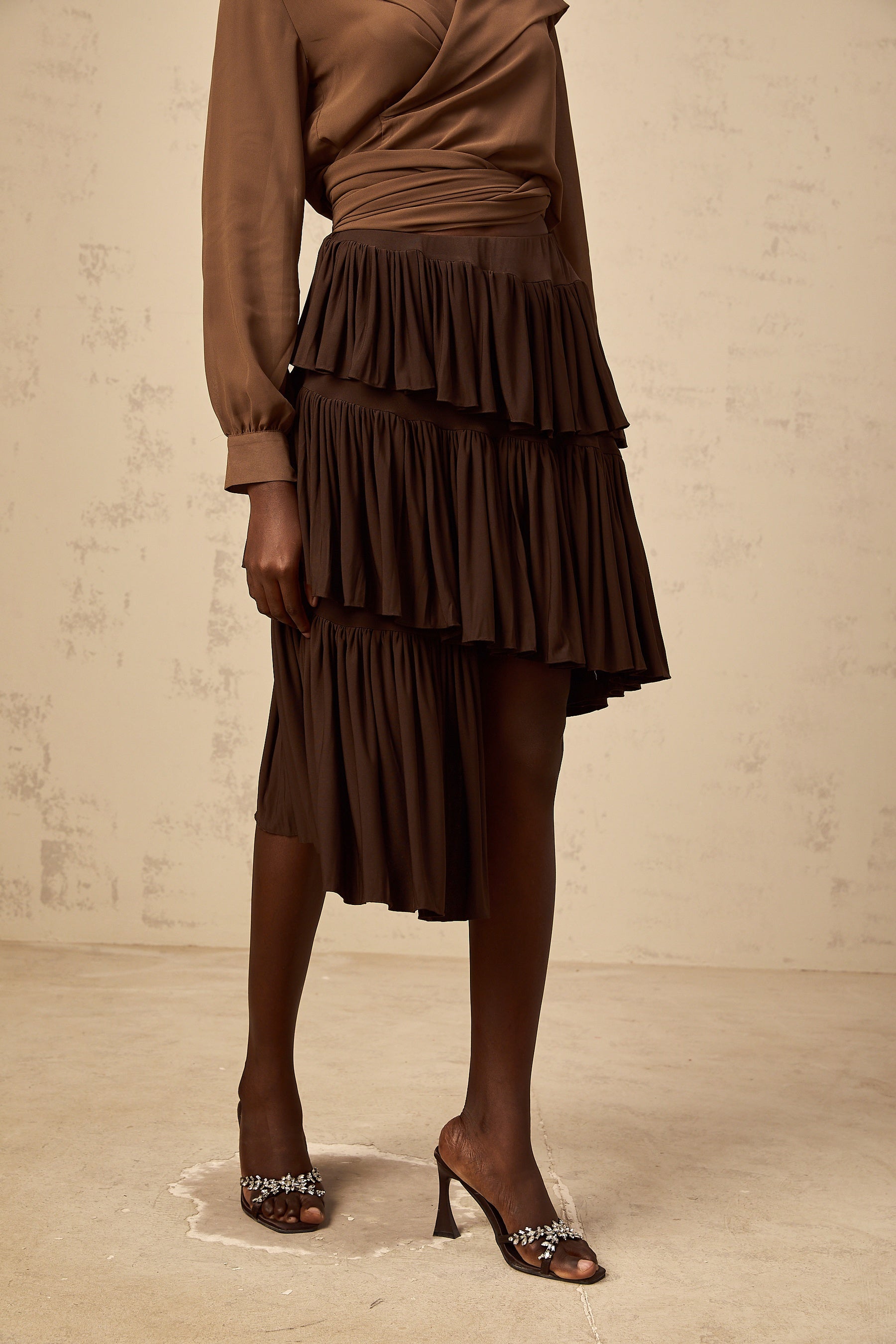 A brown asymmetrical mini skirt with pleated tiers and a unique layered design worn by a model against a plain background