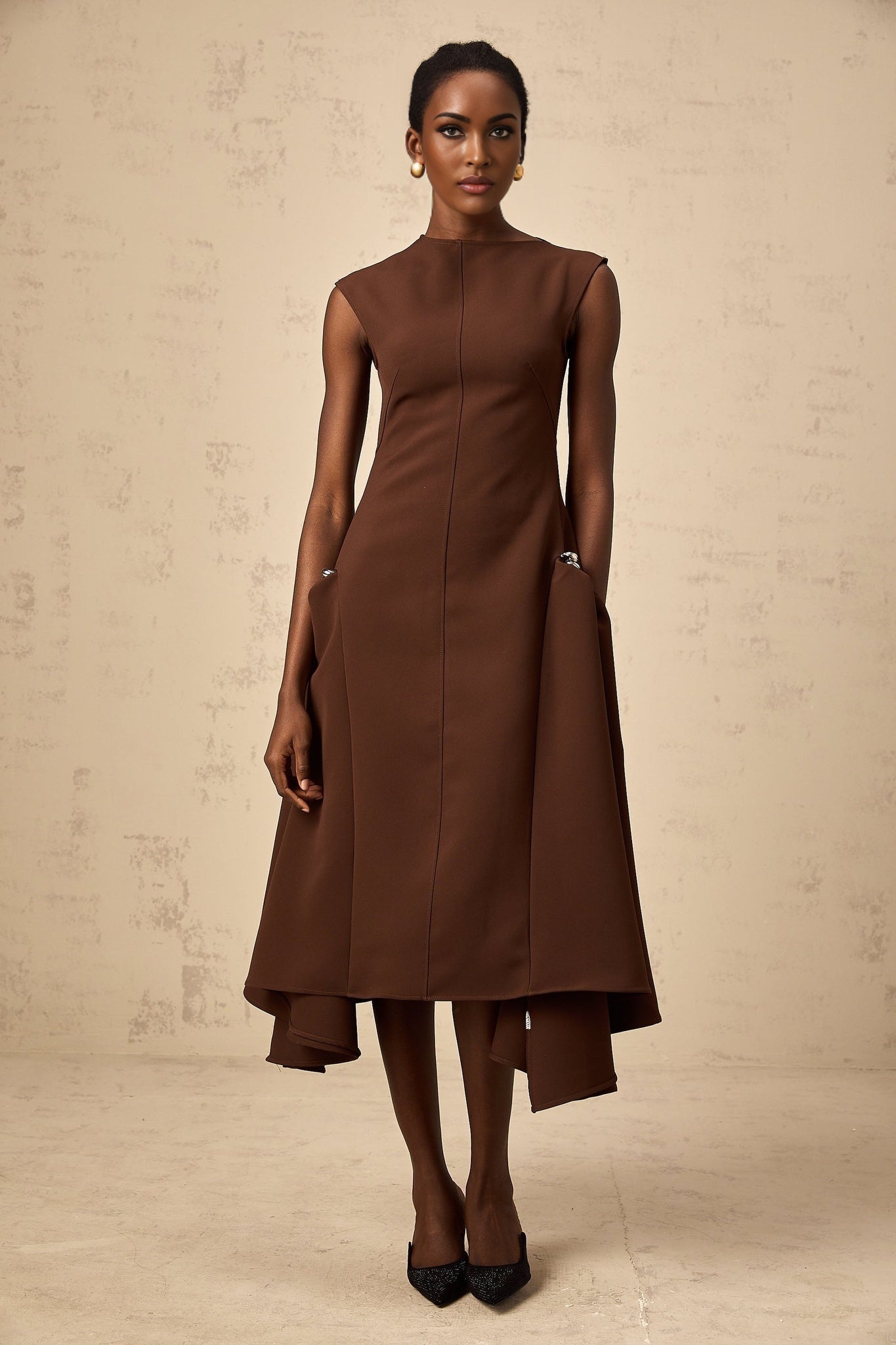 A brown midi dress with an asymmetric design worn by a model standing against a plain background