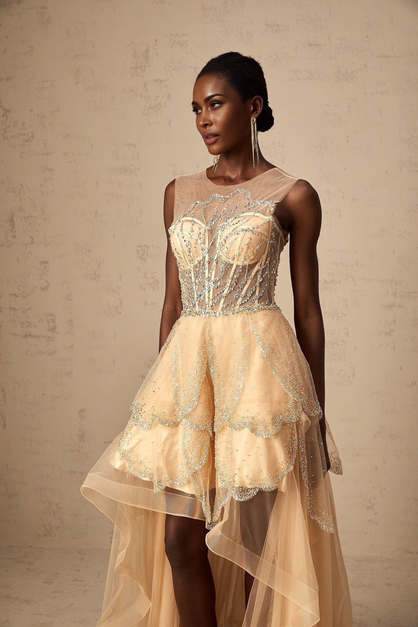A champagne colored floorlength gown with a highlow hemline featuring daisy embellishments and a flowy silhouette
