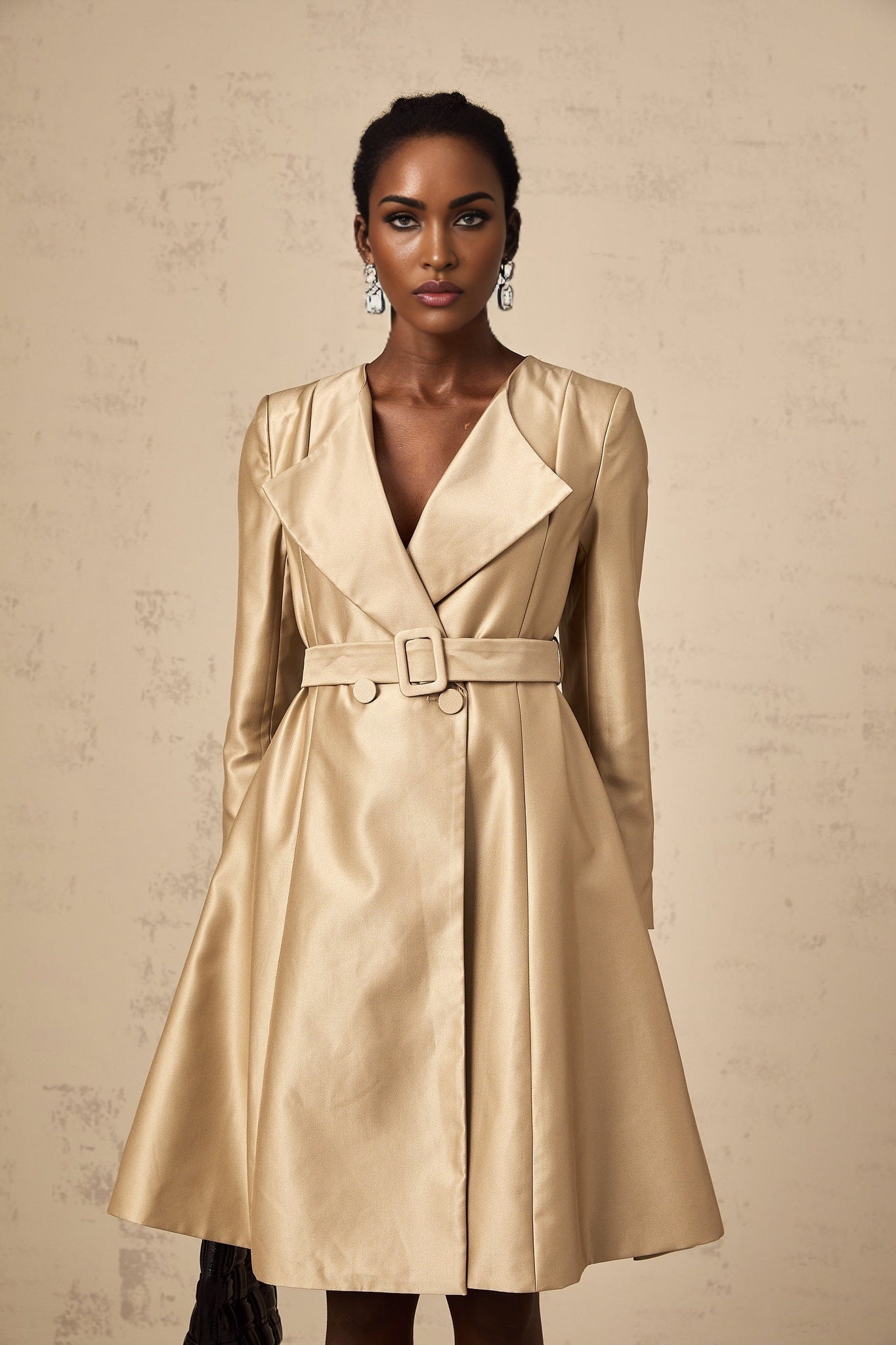 A khaki colored long coat with a belted waist worn by a model against a plain background