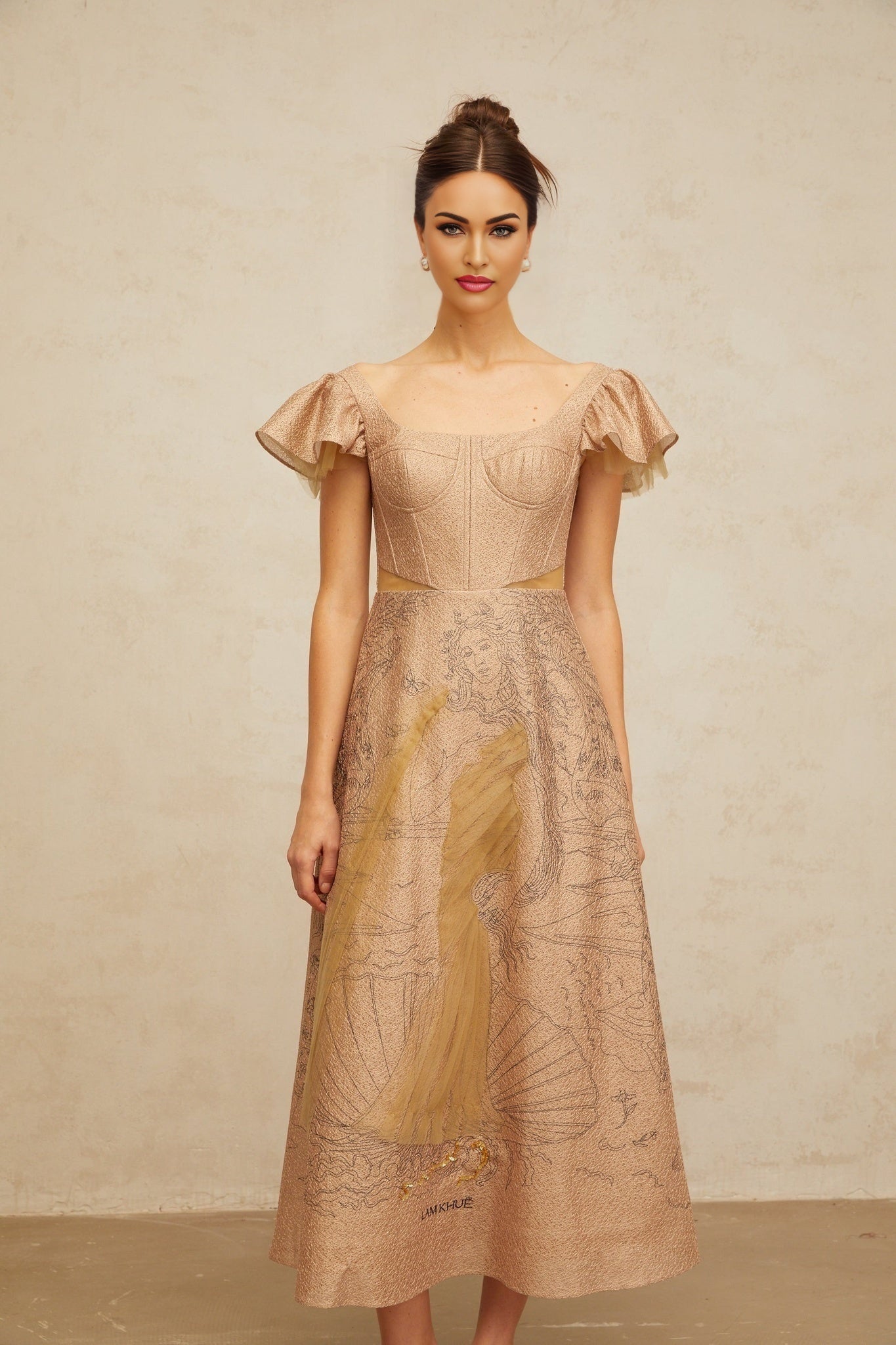 A khaki colored midi dress with a flared skirt featuring a graphic print pattern worn by a model standing in a neutral backdrop