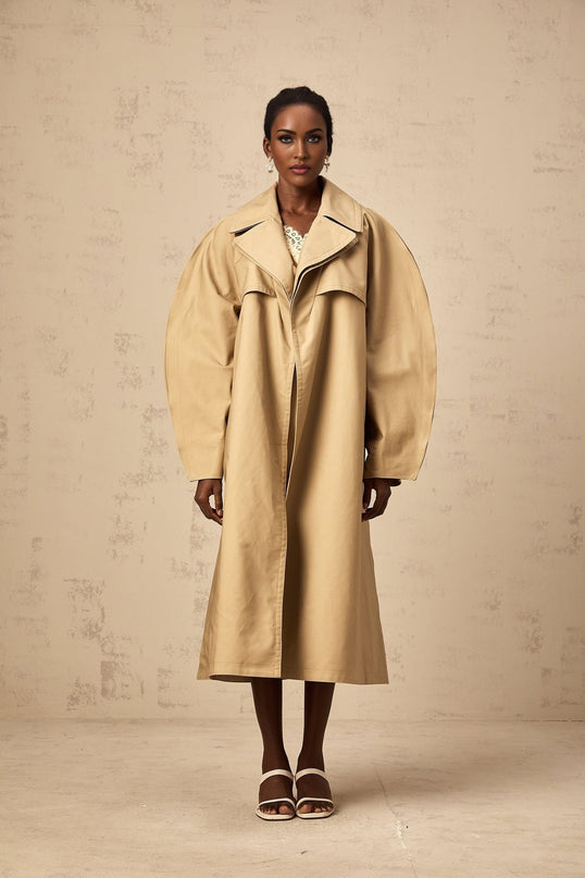 A khaki trench coat with curved sleeves worn by a model featuring a belted waist and classic lapels