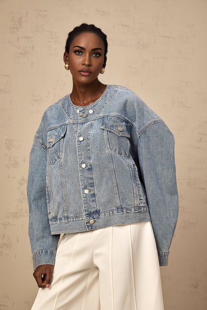 A light blue denim jacket without a collar worn by a woman against a plain background