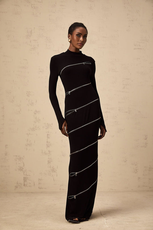 A long black fitted maxi dress with zipper details worn by a model standing against a plain background