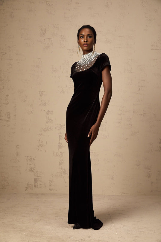 A long black velvet maxi dress with faux pearl embellishments worn by a model standing against a plain background