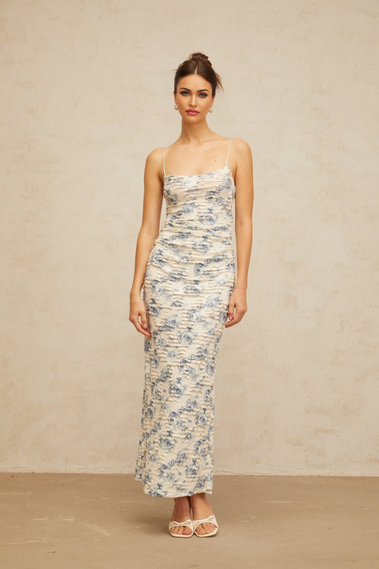 A long blue maxi dress with a textured floral print pattern covering the entire garment The dress features a flowing silhouette and appears to be made of lightweight fabric suitable for warm weather