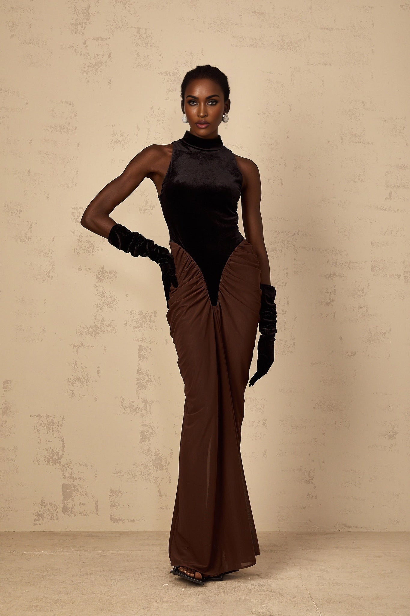 A long brown dress with velvet panels and ruched detailing named Mathilde standing on a plain background