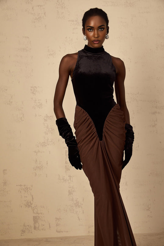 A long brown dress with velvet panels and ruched detailing named Mathilde worn by a model standing against a plain background