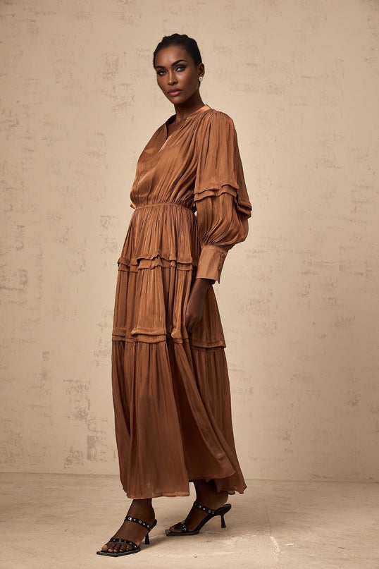 A long brown maxi dress with ruffled trim and tiered layers worn by a model standing in a neutral setting