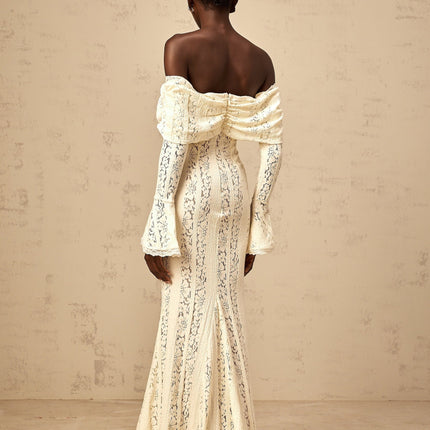 A long neutral colored maxi dress with floral lace detailing and a train design worn by a model standing in a studio setting
