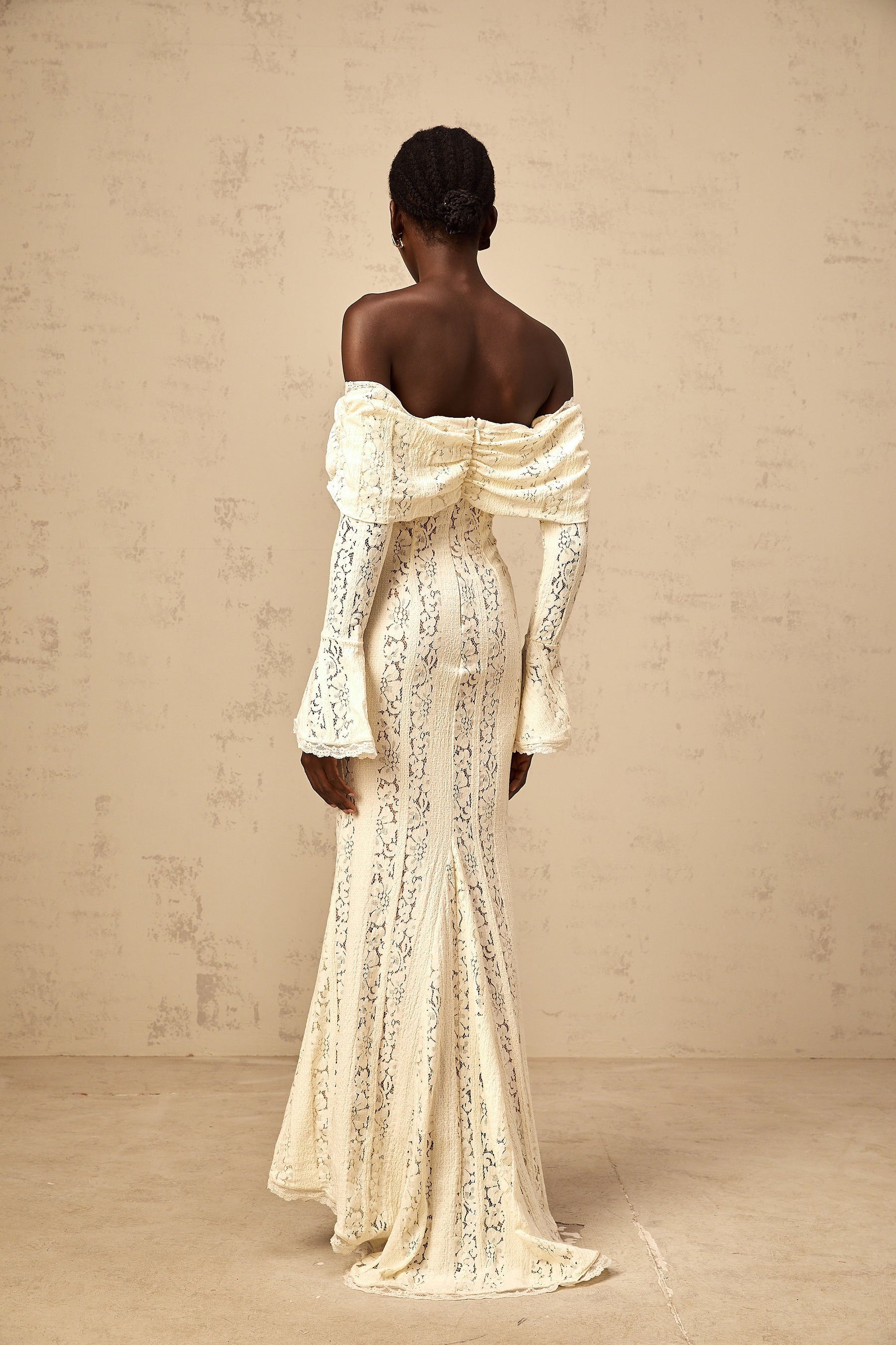 A long neutral colored maxi dress with floral lace detailing and a train design worn by a model standing in a studio setting
