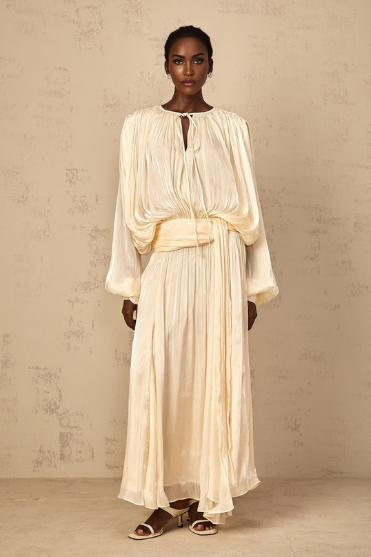 A long neutral colored maxi dress with pleated details and ruffle accents featuring a flowy silhouette and feminine design