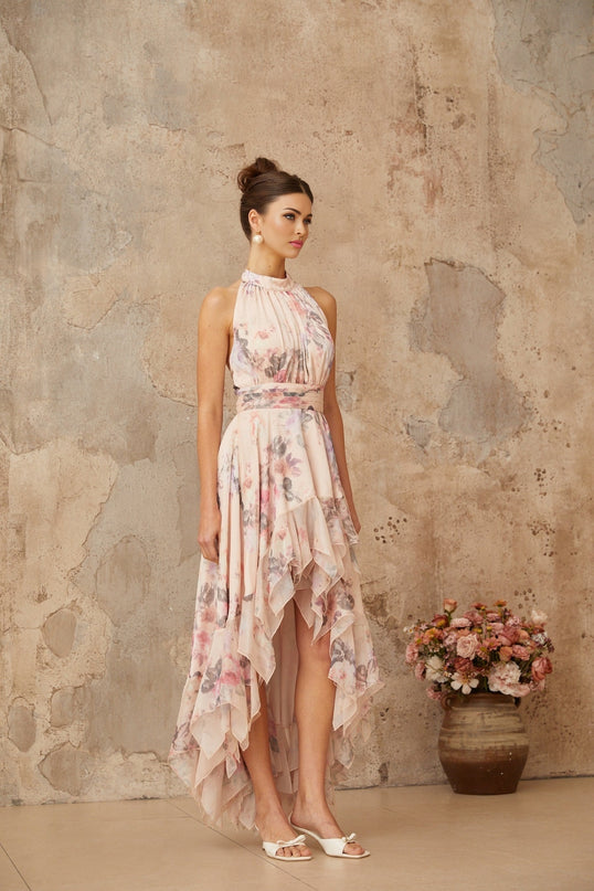 A long pink maxi dress with an asymmetrical hemline featuring a floral print pattern worn by a model against a plain background