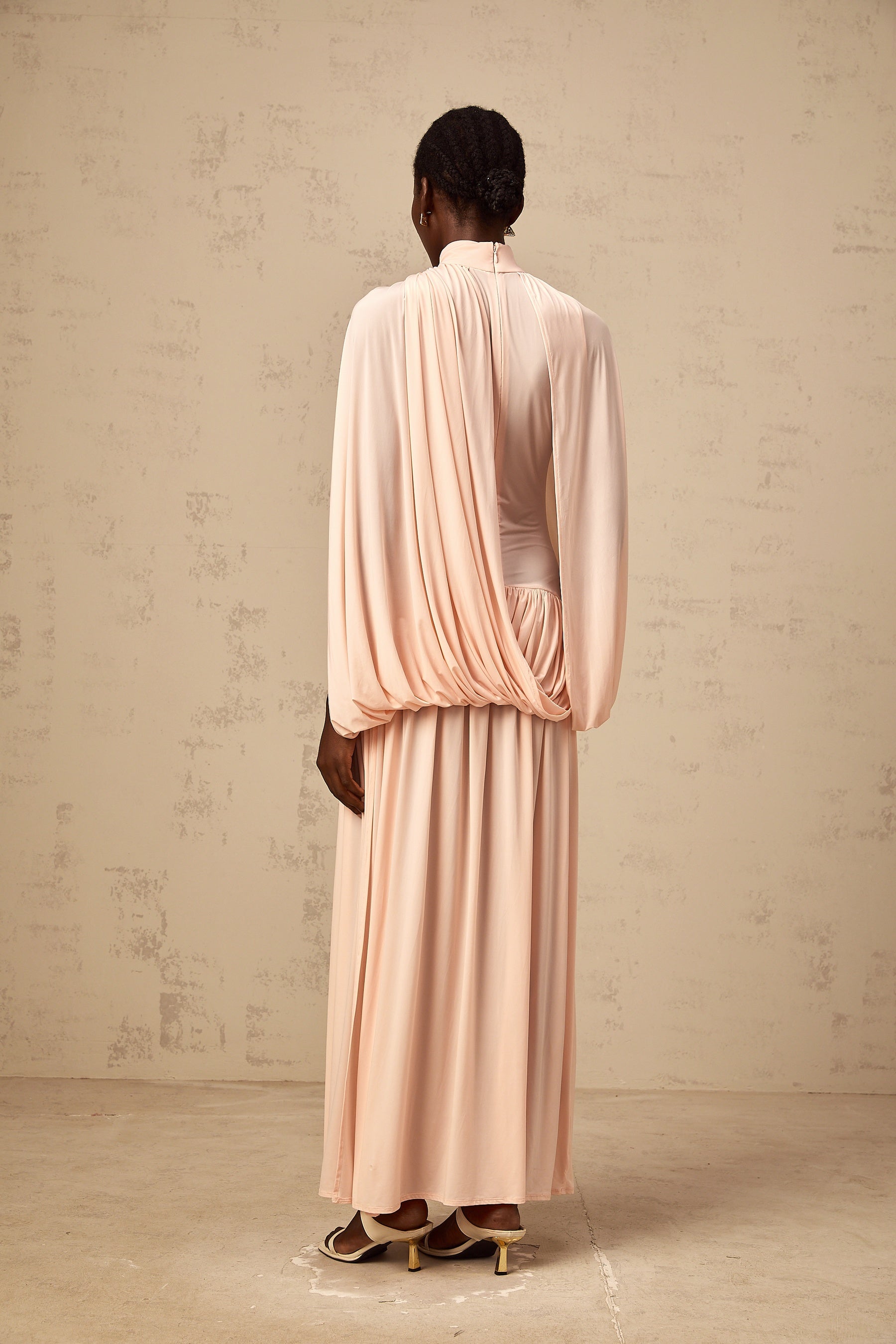 A long pink maxi dress with an asymmetrical hemline featuring a flowing cape design draped over one shoulder