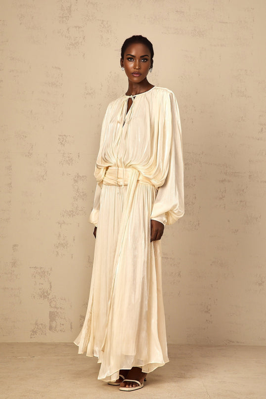 A long pleated maxi dress with ruffle details in a neutral color worn by a model standing in a casual pose