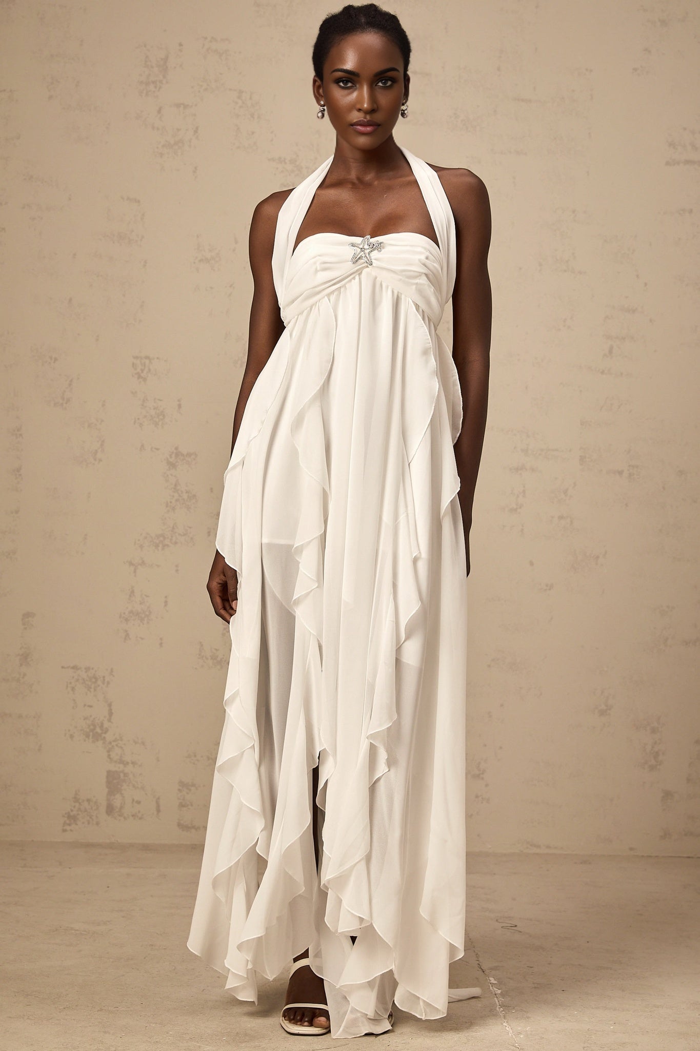 A long white chiffon maxi dress with ruffle details and a halter neckline worn by a model against a plain background