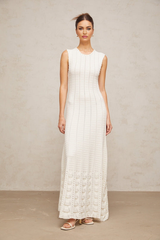 A long white knit maxi dress with a pointelle pattern and faux pearl embellishments worn by a model against a plain background