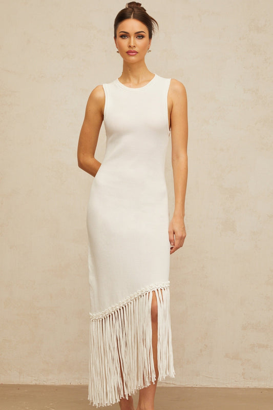 A long white knit maxi dress with fringed details worn by a model standing against a plain background