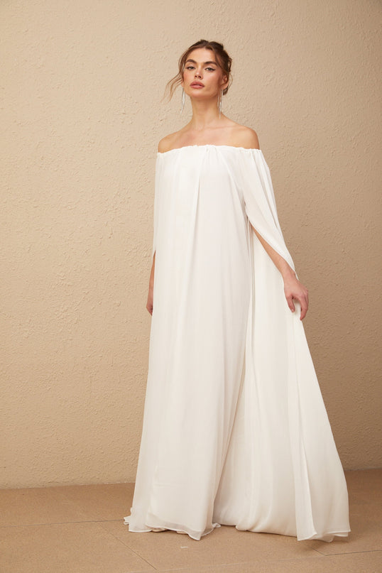 A long white maxi dress with a draped tulle design and capelike sleeves worn by a model standing in a neutral background