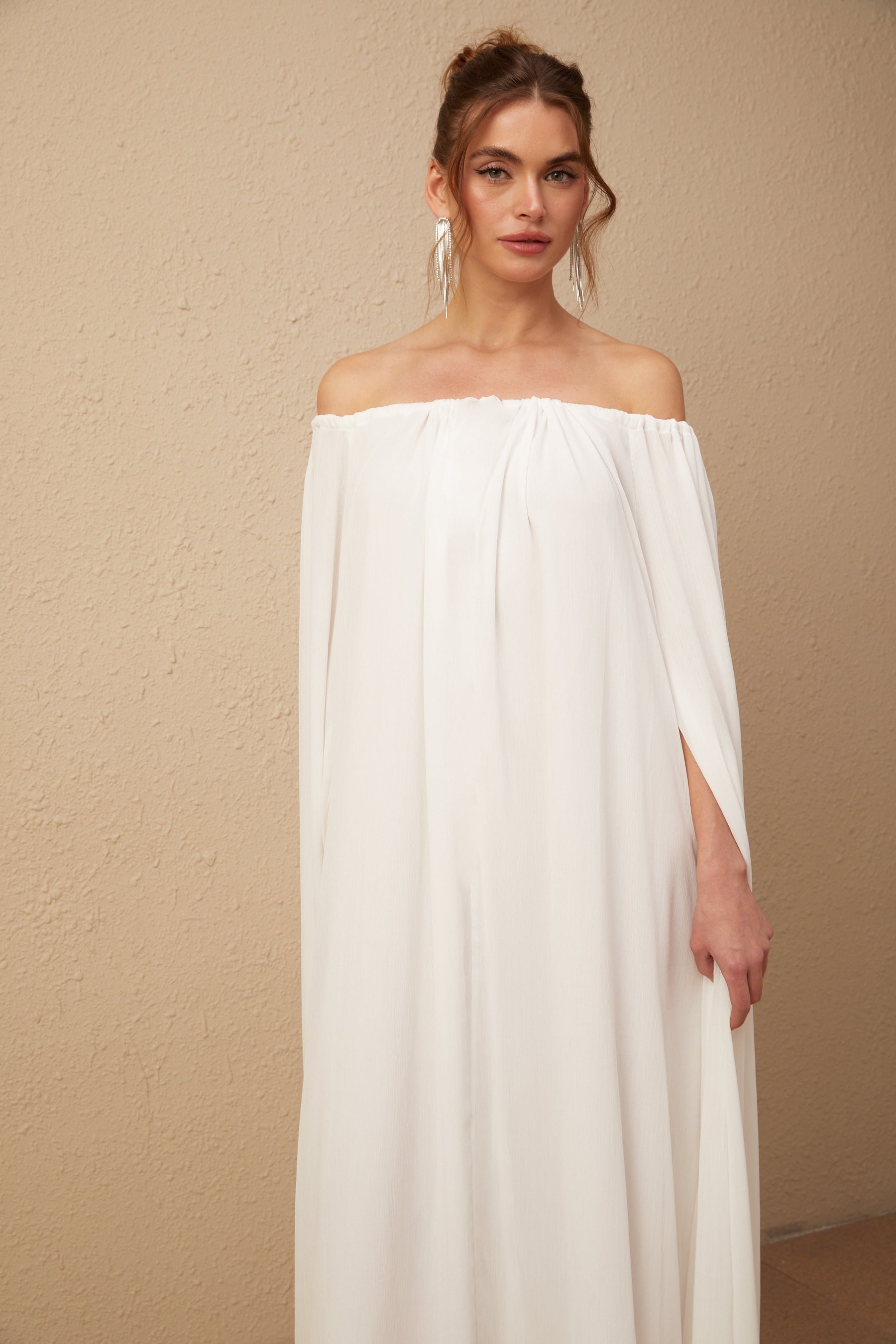 A long white maxi dress with a flowy tulle skirt and a capelike design draping over the shoulders creating an elegant silhouette