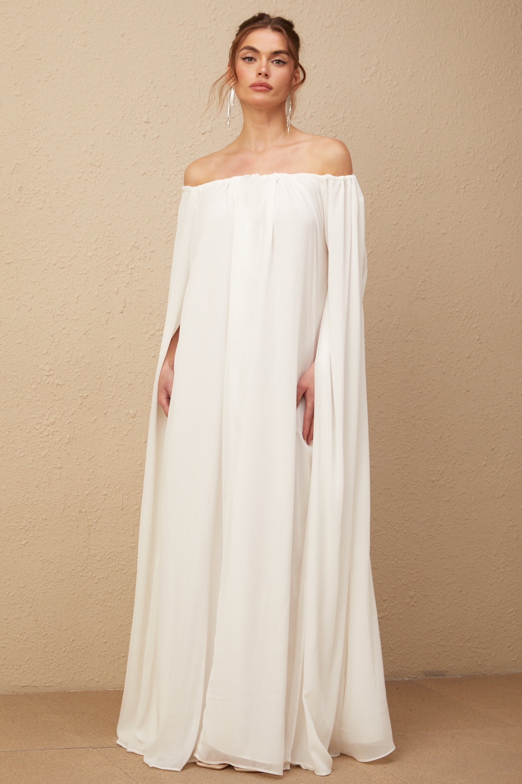 A long white tulle maxi dress with a draped cape design worn by a model standing against a plain background