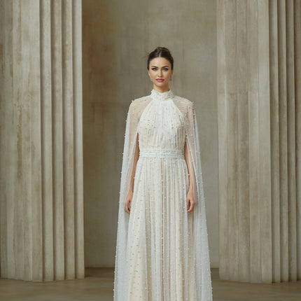 A long white tulle maxi dress with cape design and embellishments worn by a model against a plain background