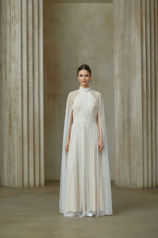 A long white tulle maxi dress with cape design and embellishments worn by a model against a plain background