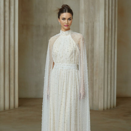 A long white tulle maxi dress with cape design and embellishments worn by a model standing against a plain background