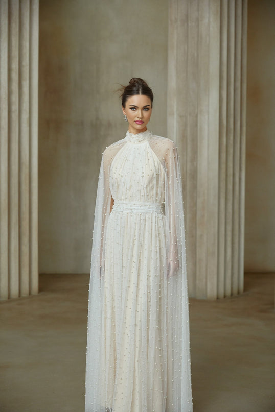 A long white tulle maxi dress with cape design and embellishments worn by a model standing against a plain background