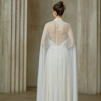 A long white tulle maxi dress with capelike design and embellishments worn by a model against a plain background