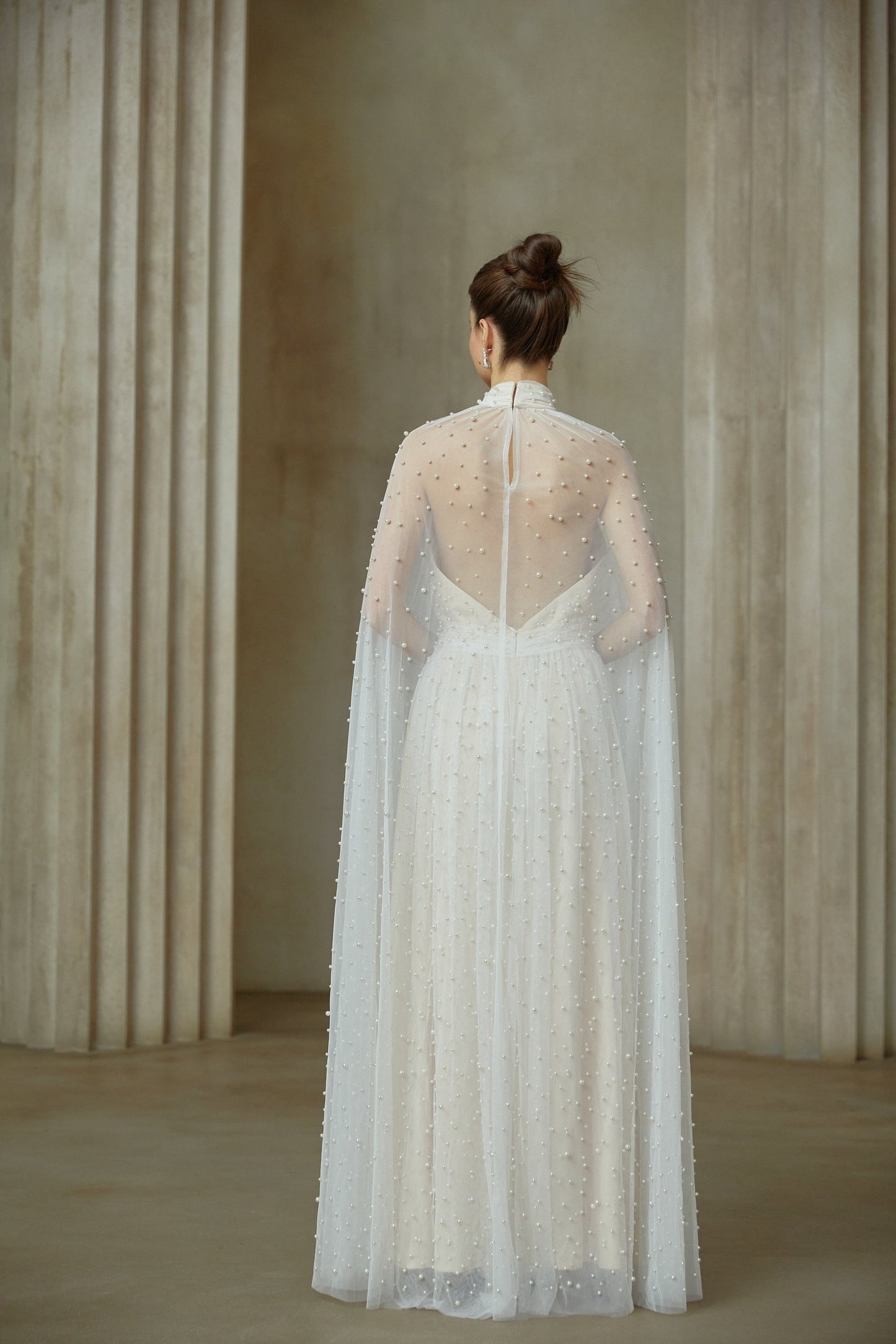 A long white tulle maxi dress with capelike design and embellishments worn by a model against a plain background
