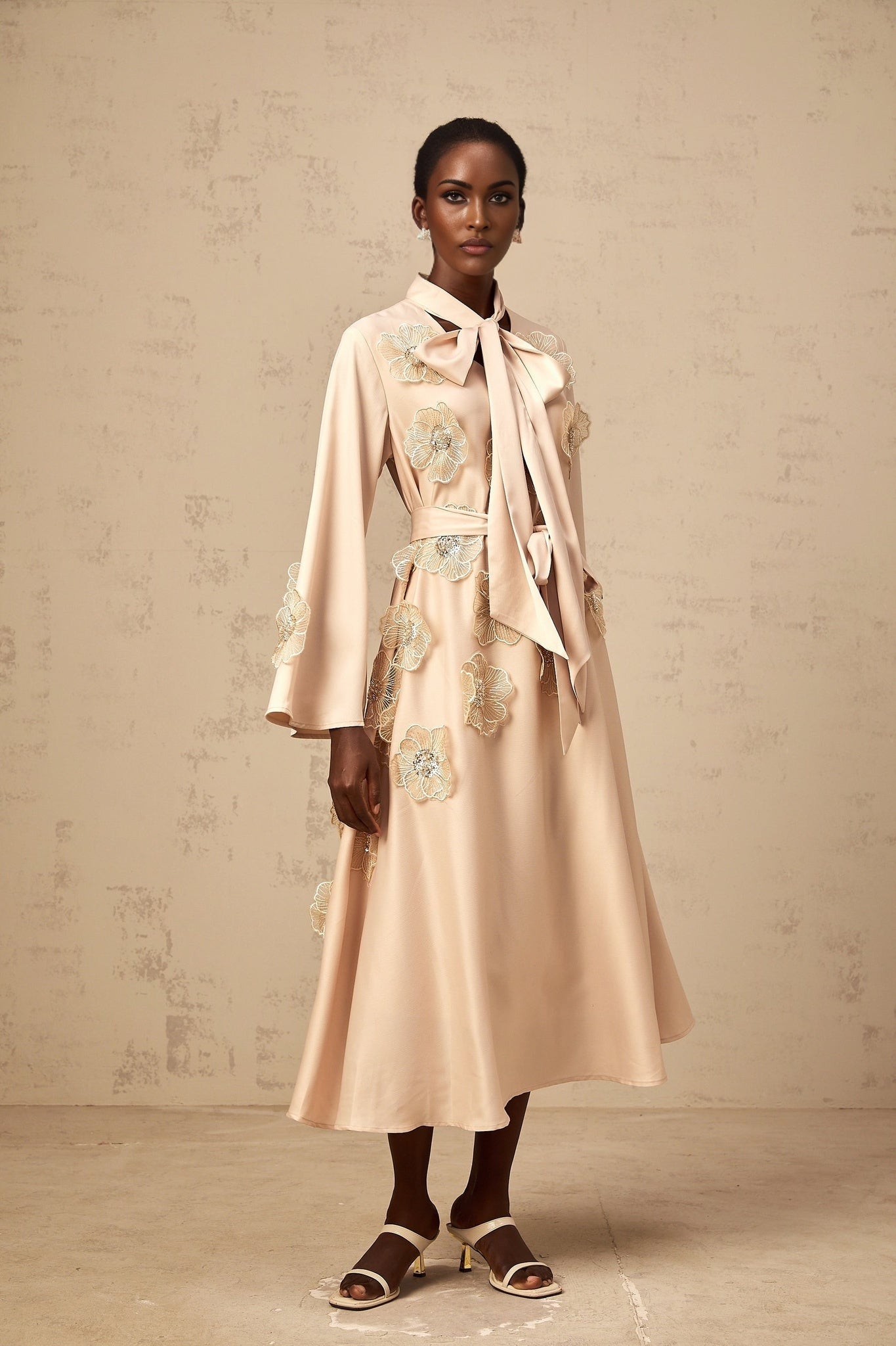 A neutralcolored flared midi dress with a tied waist and floral applique details featuring an Orane design