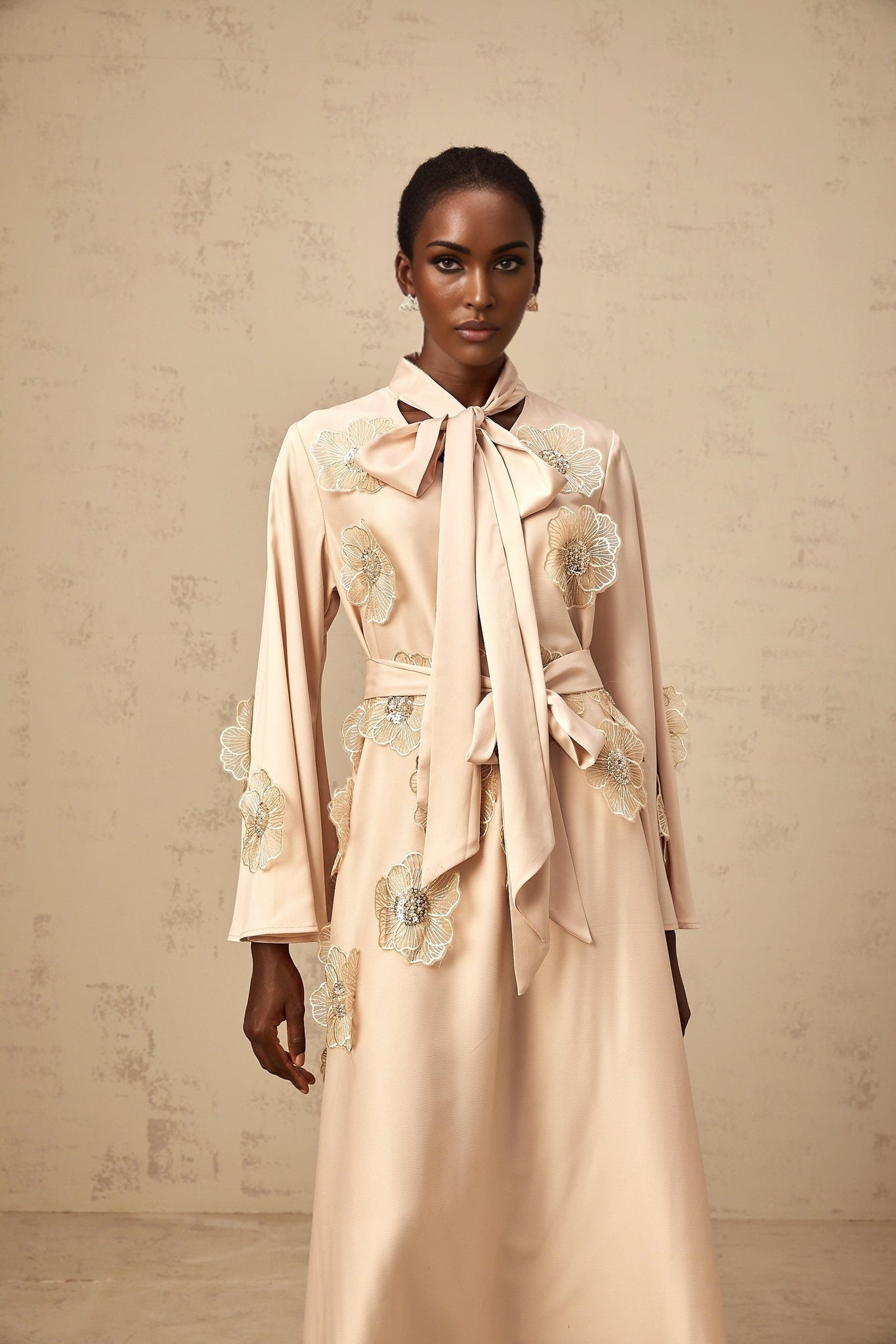 A neutralcolored flared midi dress with floral applique details and a tied waist featuring an Orane design