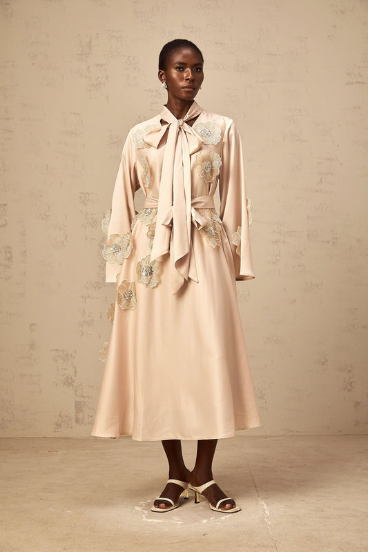 A neutralcolored flared midi dress with floral applique details and a tied waist featuring the Orane design