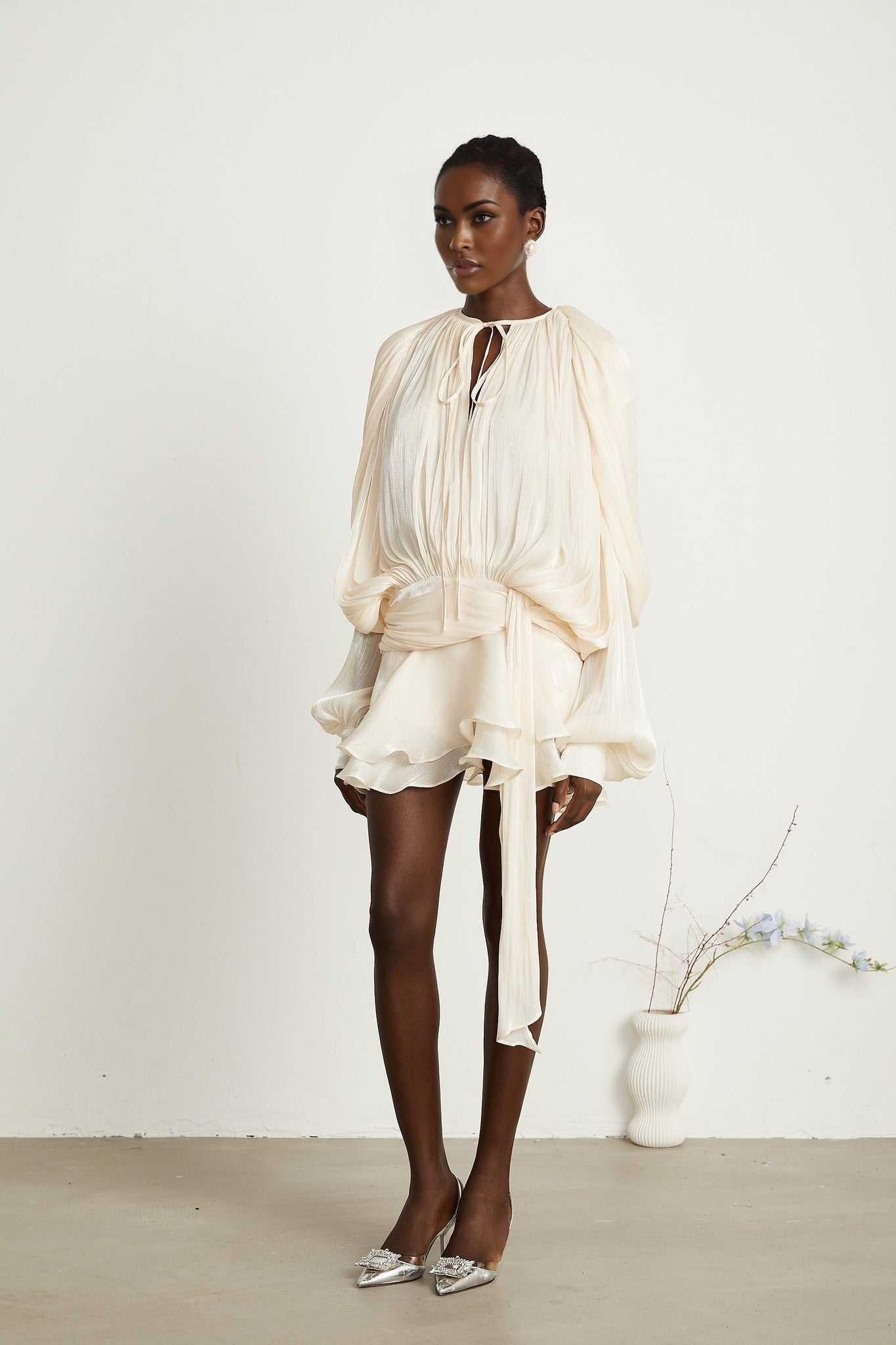 A neutralcolored short dress with a fully pleated design and ruffled details featuring a mini length and feminine silhouette