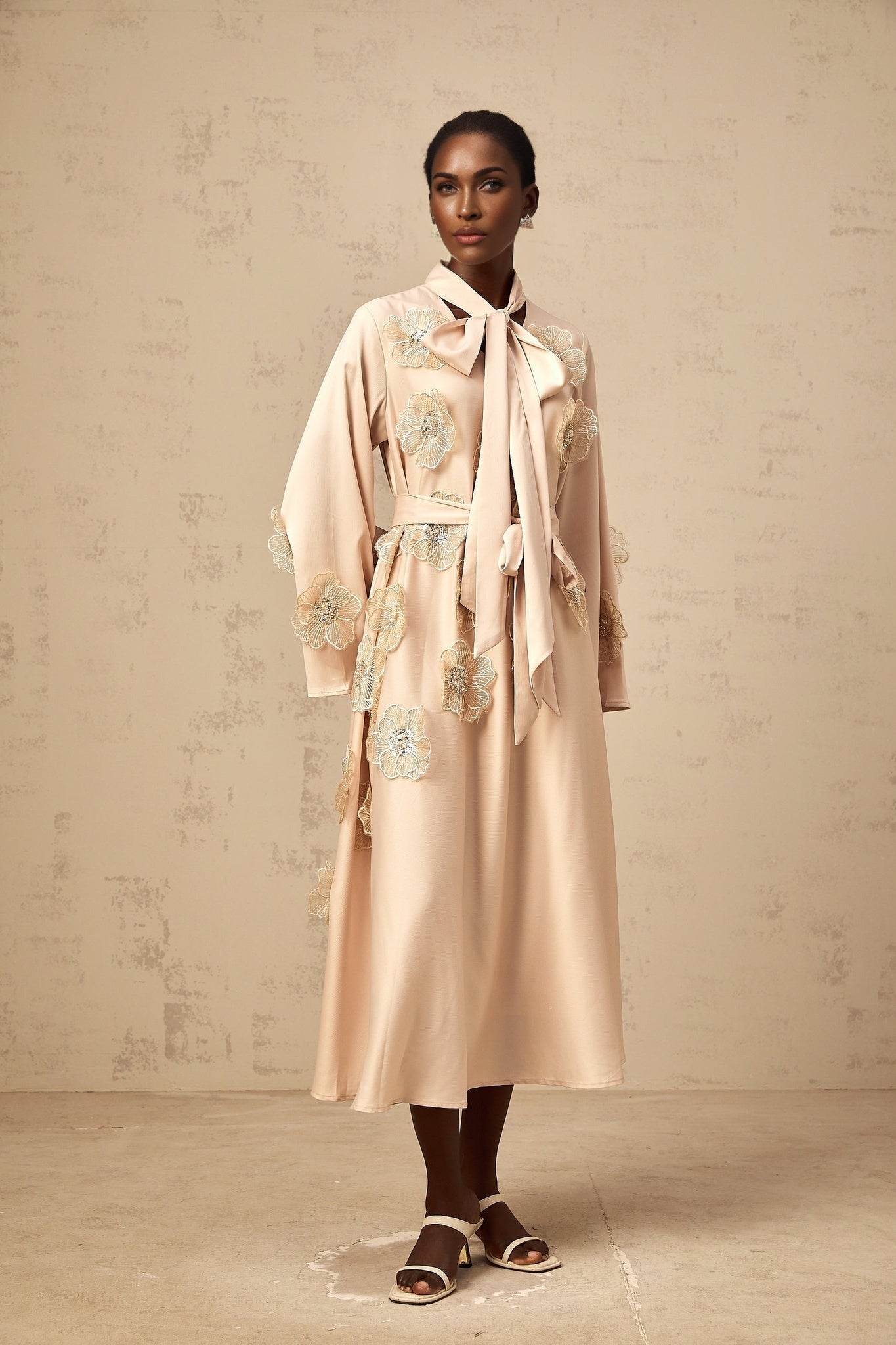 A neutraltoned flared midi dress with a tied waist and floral applique details featuring an Orane design