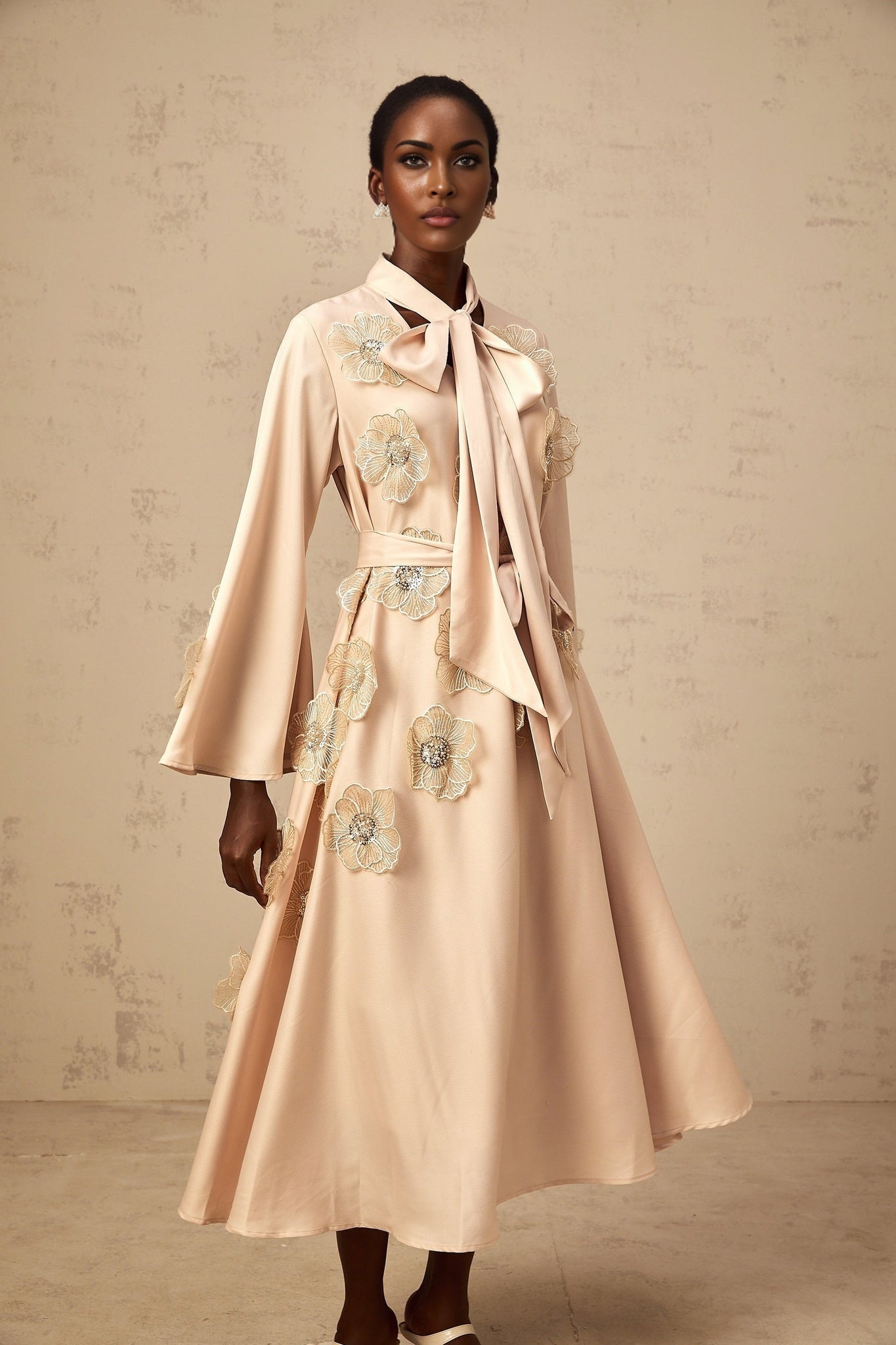 A neutraltoned midi dress with a flared silhouette featuring floral applique details and a tied waist design