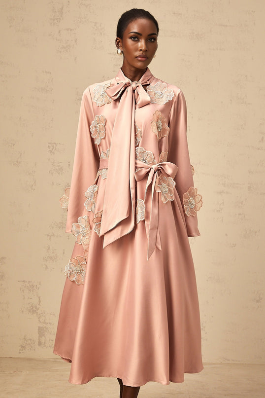 A pink flared midi dress with floral applique details and a tied waist featuring a feminine silhouette and delicate floral embellishments