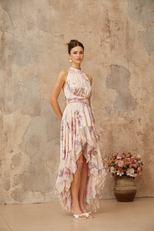 A pink maxi dress with an asymmetrical hemline featuring a floral print pattern worn by a model standing against a plain background