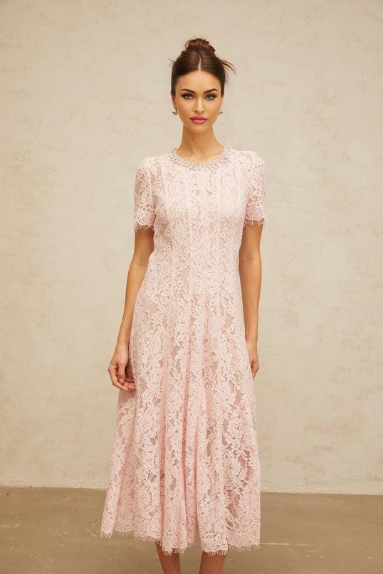 A pink midi dress with floral lace pattern and embellishments worn by a model standing against a plain background