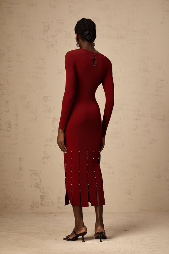 A red knit midi dress with long sleeves beaded details and strategically placed cutouts worn by a model against a plain background
