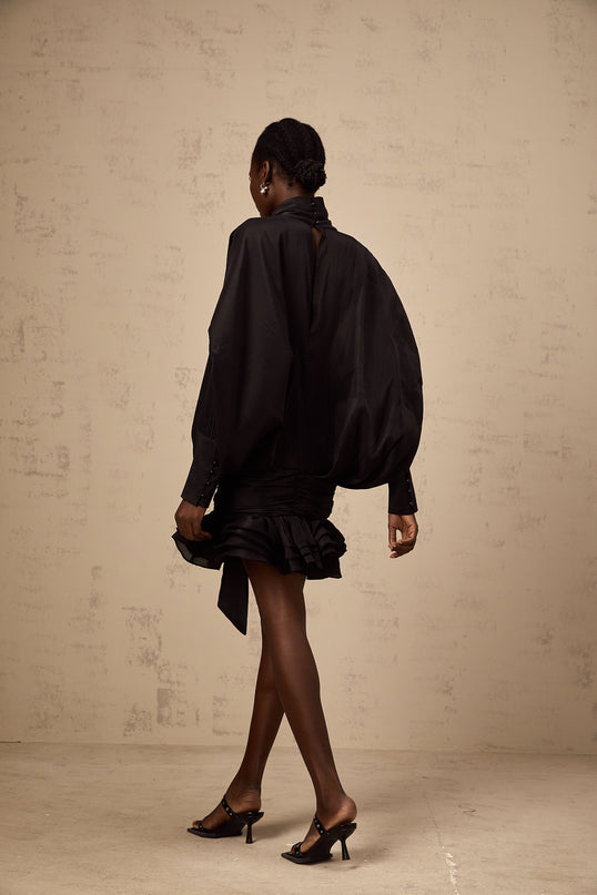 A short black mini dress with ruffled details and a pussybow neckline worn by a model against a plain background
