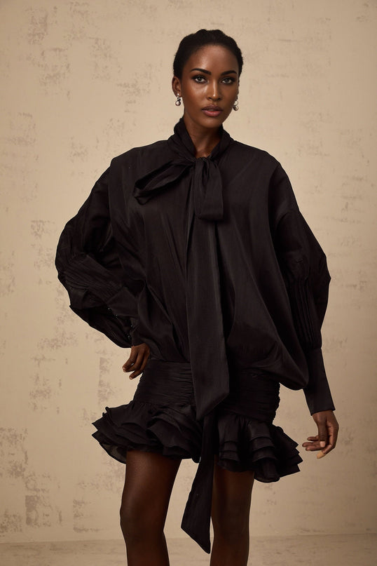 A short black mini dress with ruffled details and a pussybow neckline worn by a model named Melanie