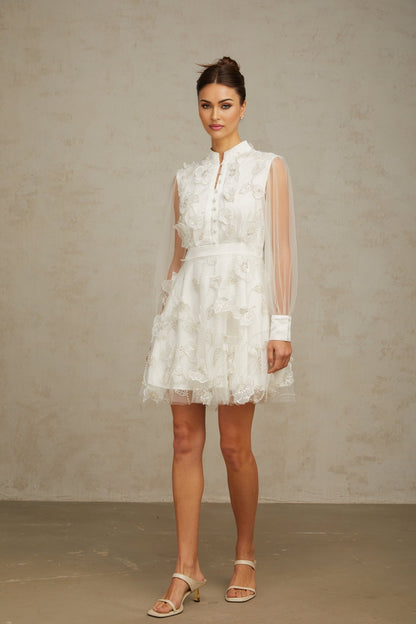 A short white mini dress with a flared skirt featuring delicate 3D tulle petals covering the bodice and scattered throughout creating a romantic floral texture