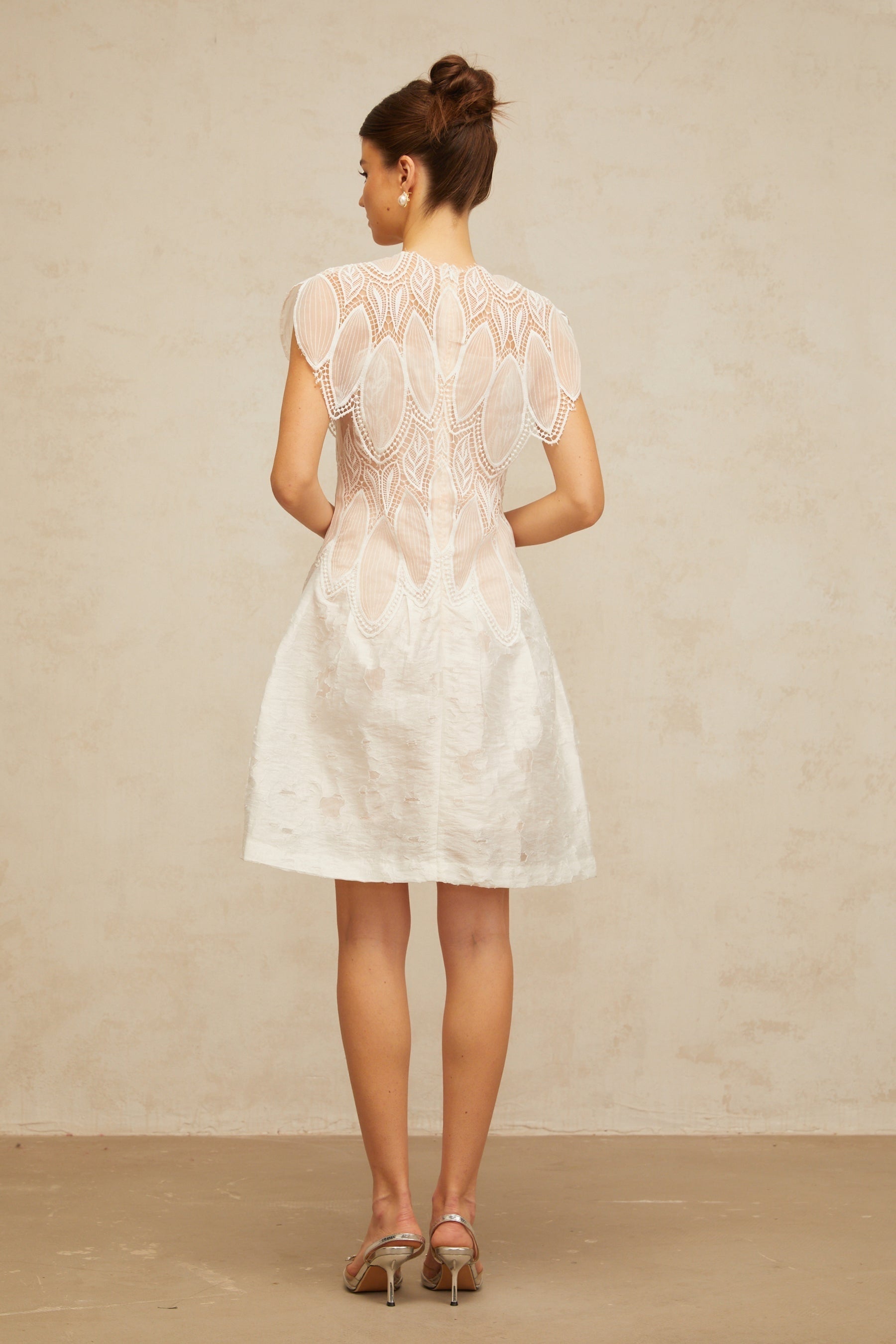 A short white mini dress with intricate 3D floral lace patterns featuring corded detailing and textured petal accents throughout the design