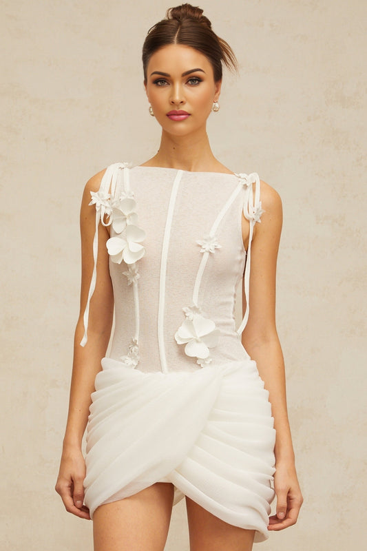 A short white mini dress with ruched fabric and floral applique details worn by a model against a plain background