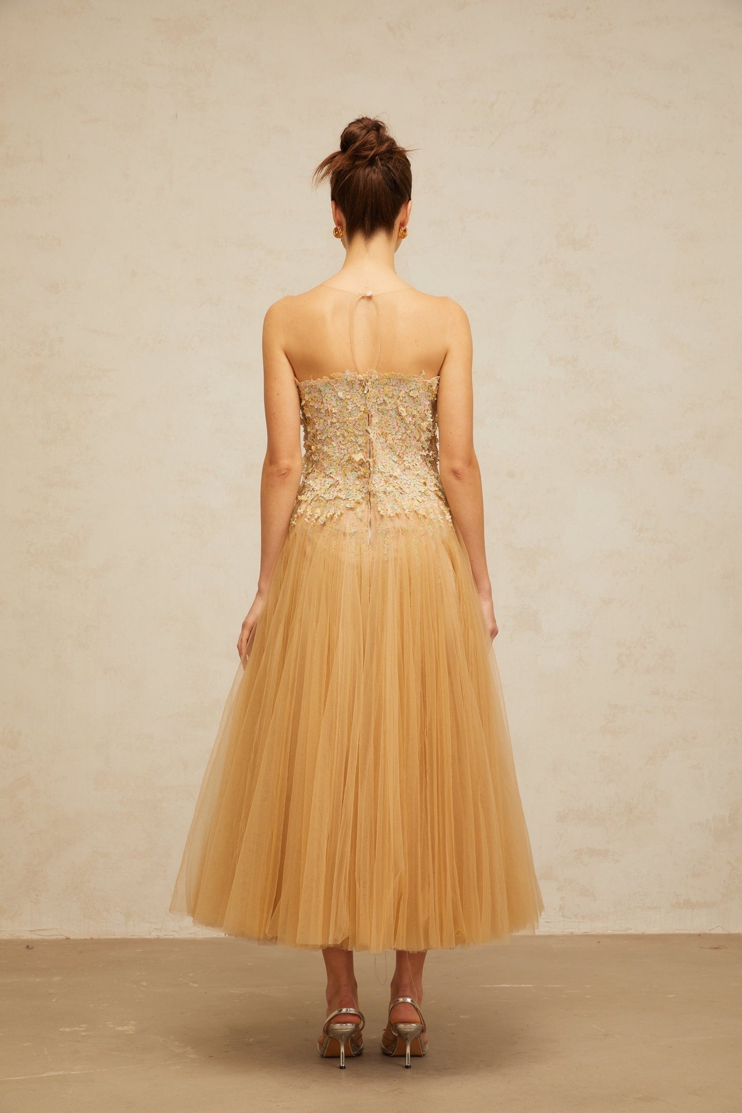 A sleeveless midi dress in neutral tones featuring pleated tulle fabric adorned with delicate sequins creating a shimmering effect throughout the garment