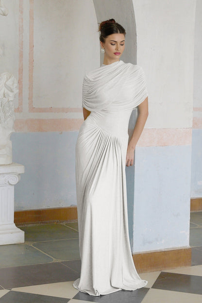 A white asymmetric pleated maxi dress with a flowing silhouette worn by a model standing against a plain background