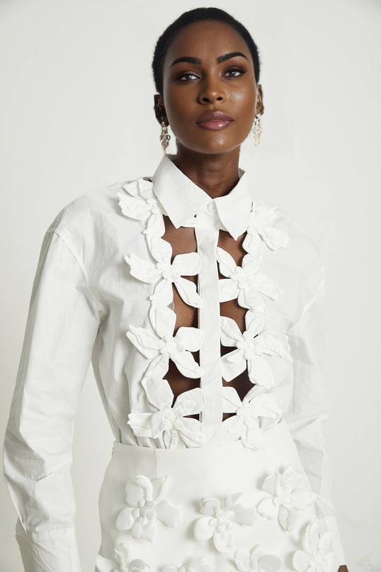 A white buttonup shirt with floral cutout patterns and short sleeves worn by a female model against a plain background
